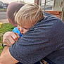 Gemma is registered to the contest to win money with this photo: adult, affection, blonde_hair, blue_clothing, brick_building, casual, child, closeup, daytime, emotional, family, grass, gray_shirt, head, hug, outdoor, person, sidewalk, skin, window