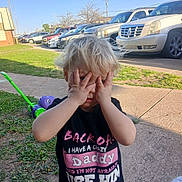 Gemma is registered to the contest to win money with this photo: blond_hair, cars, casual, child, clothing, daylight, face_covering, grass, hands, lawnmower, outdoor, parking_lot, person, playful, shirt, sidewalk, text, timestamp, toddler, toy