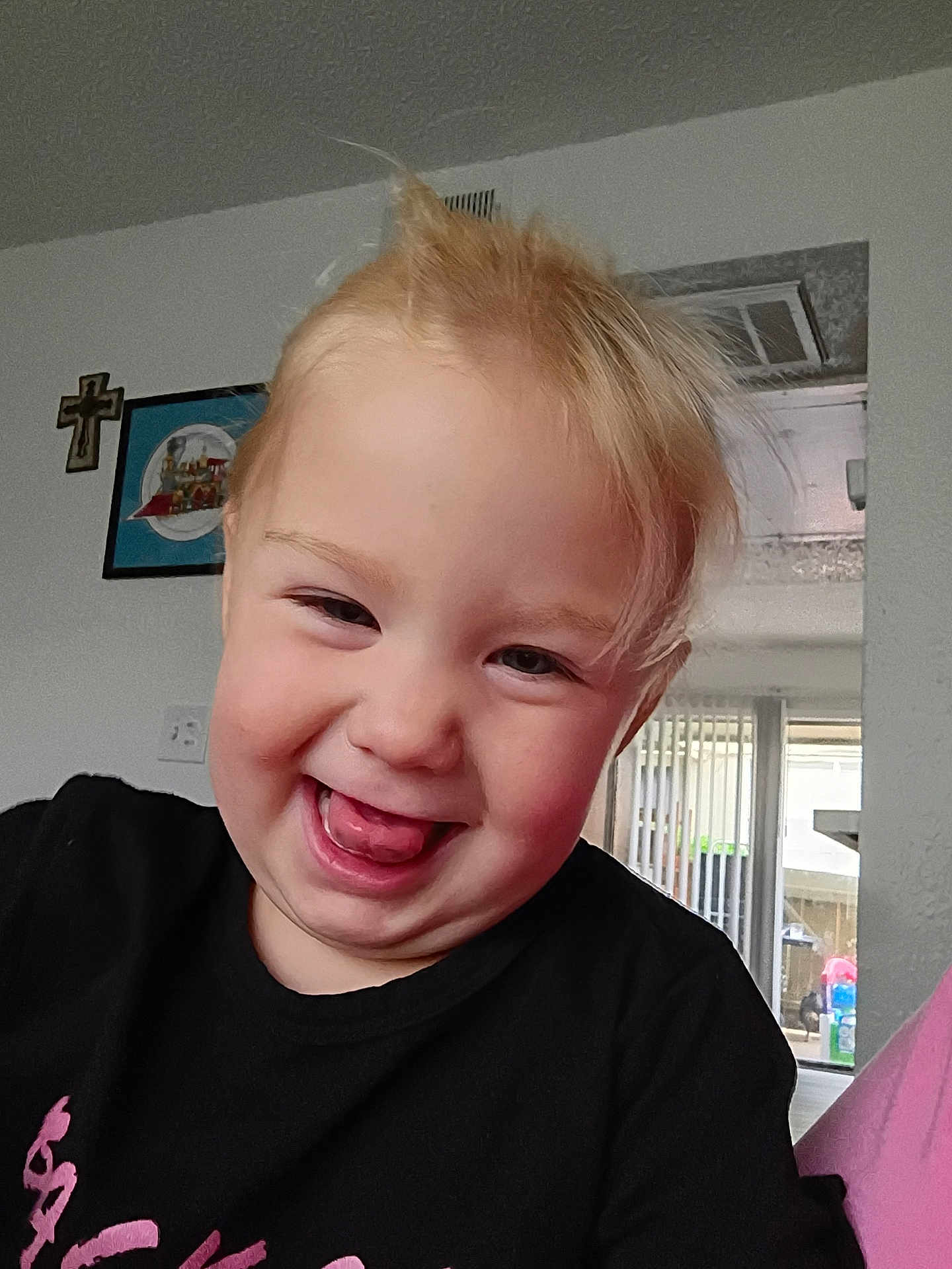 Gemma joined the competition — help win amazing prizes! toddler, child, smiling, tongue_out, blonde_hair, black_shirt, indoor, home, wall_art, cross, window, natural_light, happy, playful, portrait, face, person, cute, young_child, casual