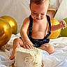 baby, cake, birthday, messy, celebration, white_cake, cream, golden_balloons, suspenders, child, smiling, sitting, first_birthday, food, happy, face, hand, party, indoors, cute
