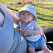 Cody is registered to the contest to win money with this photo: baby, infant, bucket_hat, onesie, sitting, lap, parent_arm, hand, bracelet, hope_tattoo, park, grass, bench, pond, sunlight, shadow, portrait, expression, outdoor, casual_clothing