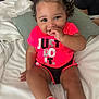 Skylar joined the competition — help win amazing prizes! toddler, child, curly_hair, pink_shirt, shorts, sitting, bed, pillow, white_bedding, smiling, hand_near_mouth, hair_ties, bright_colors, indoor, person, cute, happy, portrait, footwear, socks