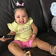 Skylar is registered to the contest to win money with this photo: toddler, child, smiling, tongue_out, curly_hair, pink_bow, yellow_shirt, pink_shorts, sandals, remote_control, leather_couch, sitting, indoor, bracelets, happy, person, clothing, footwear, cute, playful