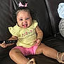 toddler, child, smiling, tongue_out, curly_hair, pink_bow, yellow_shirt, pink_shorts, sandals, remote_control, leather_couch, sitting, indoor, bracelets, happy, person, clothing, footwear, cute, playful