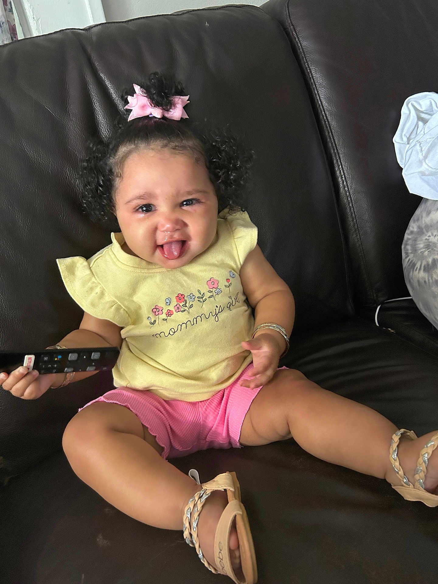 Skylar is registered to the contest to win money with this photo: toddler, child, smiling, tongue_out, curly_hair, pink_bow, yellow_shirt, pink_shorts, sandals, remote_control, leather_couch, sitting, indoor, bracelets, happy, person, clothing, footwear, cute, playful