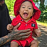 baby, child, red_clothing, red_hat, laughing, tattooed_hand, adult, outdoor, trees, sunlight, park, greenery, smile, happy, person, casual_clothing, nature, daylight, sitting, holding