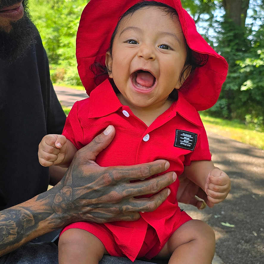 Ricardo Jasiel is registered to the contest to win money with this photo: baby, child, red_clothing, red_hat, laughing, tattooed_hand, adult, outdoor, trees, sunlight, park, greenery, smile, happy, person, casual_clothing, nature, daylight, sitting, holding