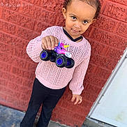 Gracie joined the competition — help win amazing prizes! child, girl, toy, monster_truck, unicorn, pink_sweater, black_pants, brown_shoes, braided_hair, brick_wall, outdoor, sidewalk, curious, standing, smiling, playful, casual_clothing, portrait, daytime, urban