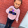 black_pants, braided_hair, brick_wall, brown_shoes, casual_clothing, child, curious, daytime, girl, monster_truck, outdoor, pink_sweater, playful, portrait, sidewalk, smiling, standing, toy, unicorn, urban