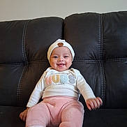 Raquel is registered to the contest to win money with this photo: baby, child, clothing, couch, cozy, cute, happy, hat, headwear, home, indoor, infant, leather, person, pink_pants, portrait, sitting, smiling, white_shirt, young_child