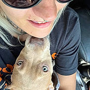 Leia is registered to the contest to win money with this photo: person, dog, puppy, face, smile, sunglasses, nose_ring, black_shirt, light_hair, car_seat, closeup, pet, affection, animal, young_dog, indoor, casual, human, cute, companion
