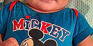 Johnnie is registered to the contest to win money with this photo: baby, blue_shirt, cartoon_character, child, clothing, colorful, cute, face, hand, happy, indoors, infant, lying_down, mickey_mouse, patterned_background, person, play_mat, short_sleeves, smiling, young_child