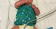 Waylon is registered to the contest to win money with this photo: baby, bed, blue_sock, cable, cuff, cute, diaper, green_shirt, healthcare, hospital, indoors, infant, lying_down, medical, medicine, newborn, patient, person, red_cuff, sock