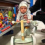 baby, child, toy_store, tricycle, wooden_toy, hand, beanie, hoodie, floor, shelf, toys, packaging, person, indoor, clothing, support, wheel, play, cute, expression