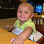 baby, child, smiling, table, highchair, indoor, restaurant, wooden_table, yellow_shirt, necklace, blanket, face, hand, cute, infant, person, furniture, dining, casual, light