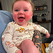 Skyler is registered to the contest to win money with this photo: baby, smiling, child, tattoo, arm, clothing, indoor, kitchen, furniture, table, person, face, happy, sitting, skin, expression, long_sleeve, background, home, infant