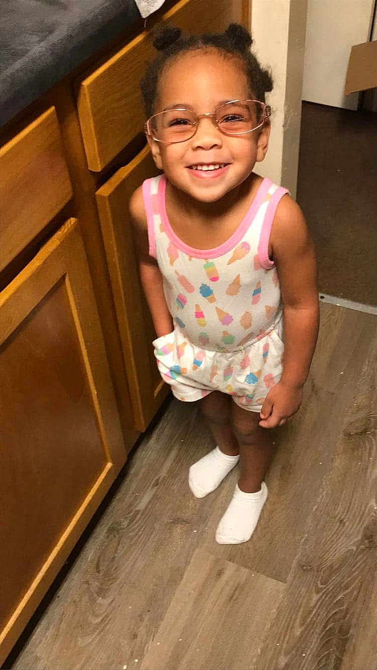 Kamarie is registered to the contest to win money with this photo: child, smiling, glasses, hair_buns, tank_top, shorts, socks, kitchen, wooden_cabinets, flooring, indoors, happy, person, cute, standing, young_child, patterned_clothing, playful, bright, casual