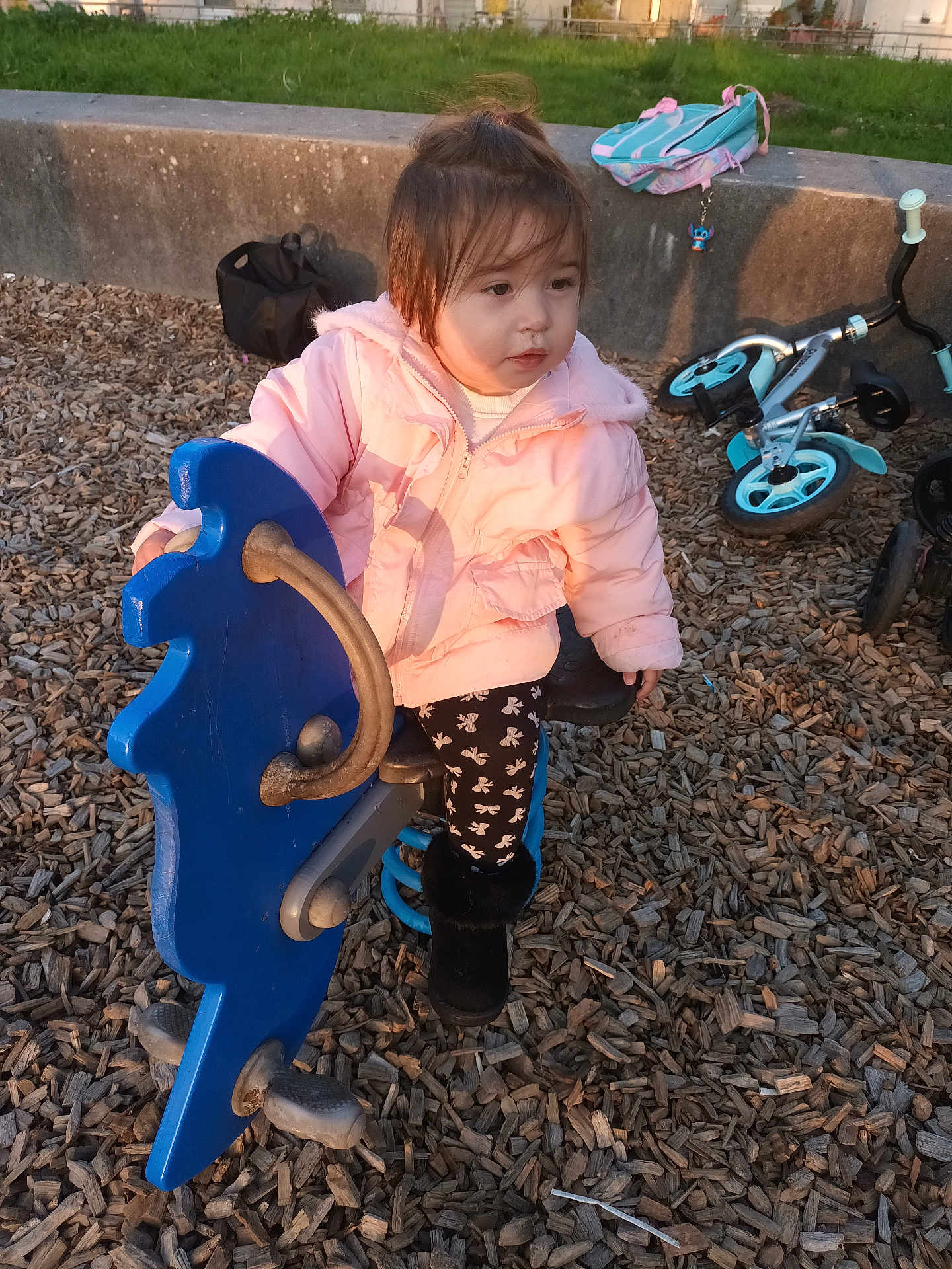 Alya a rejoint le concours — aidez-le/la à gagner de superbes lots ! toddler, child, playground, spring_rocker, blue_horse, pink_jacket, black_boots, wood_chips, bicycle, backpack, concrete_ledge, outdoor, daylight, casual_clothing, young_child, play_equipment, park, person, nature, fun