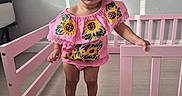 Annalise is registered to the contest to win money with this photo: child, toddler, girl, baby, portrait, standing, smiling, indoors, playpen, pink, bow, hair_accessory, sunflower, floral_clothing, dress, nursery, home, wooden_floor, furniture, photography