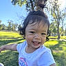 Annalise is registered to the contest to win money with this photo: child, toddler, smiling, outdoor, park, portrait, face, happy, sunlight, grass, trees, nature, playful, topknot, dark_hair, white_shirt, cartoon_shirt, teeth, candid, daylight