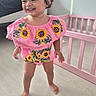 Annalise is registered to the contest to win money with this photo: child, toddler, smiling, laughing, standing, portrait, pink_outfit, floral_clothing, sunflower_pattern, hair_bow, curly_hair, bedroom, crib, nursery, wooden_floor, indoor, daylight, happy, barefoot, baby