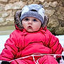 baby, infant, bundled_up, pink_snowsuit, knit_hat, fur_trim, blue_eyes, rosy_cheeks, snow, outdoor, stone_wall, mitten, winter_clothing, sitting, rope, cold_weather, portrait, puffy_jacket, cute_expression, hat_with_ears