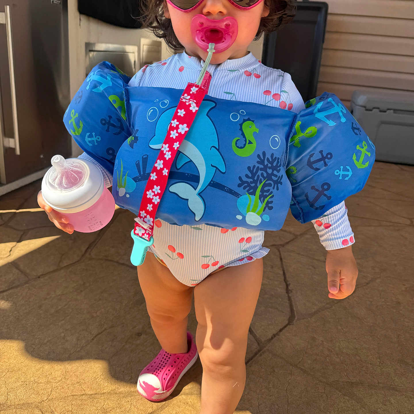 Lina is registered to the contest to win money with this photo: bottle, child, clip, concrete_floor, dolphin, floatation_vest, life_jacket, outdoor, pacifier, person, pink, sea_creatures, seahorse, shoes, standing, summer, sunglasses, sunny, swimsuit, toddler