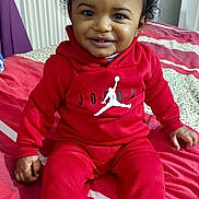 Esaia participe au concours pour gagner de l'argent avec cette photo : toddler, child, smiling, red_clothing, socks, bed, blanket, indoor, portrait, happy, cute, hair_tie, sitting, young_child, person, face, cozy, home, casual, fashion