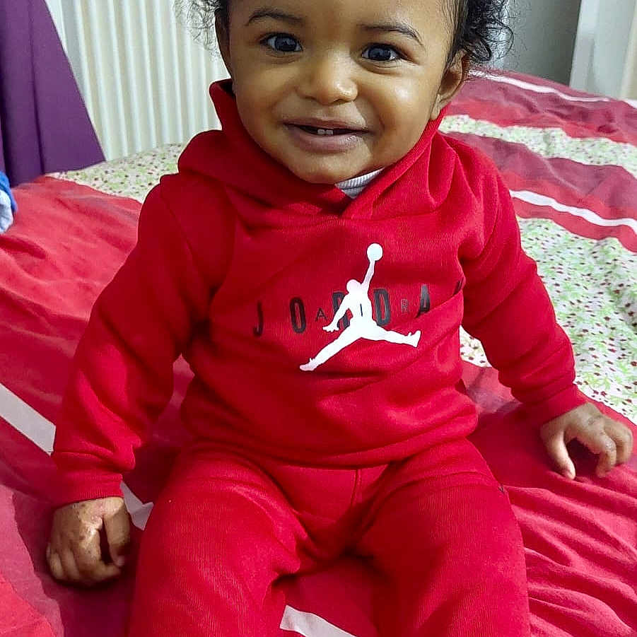 Esaia participe au concours pour gagner de l'argent avec cette photo : bed, blanket, casual, child, cozy, cute, face, fashion, hair_tie, happy, home, indoor, person, portrait, red_clothing, sitting, smiling, socks, toddler, young_child