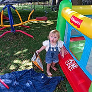 Charley is registered to the contest to win money with this photo: child, toddler, grass, playground, inflatable, snack_bag, barefoot, towel, outdoor, toy, play_structure, tree, fence, summer, sunlight, clothing, face, messy, person, nature