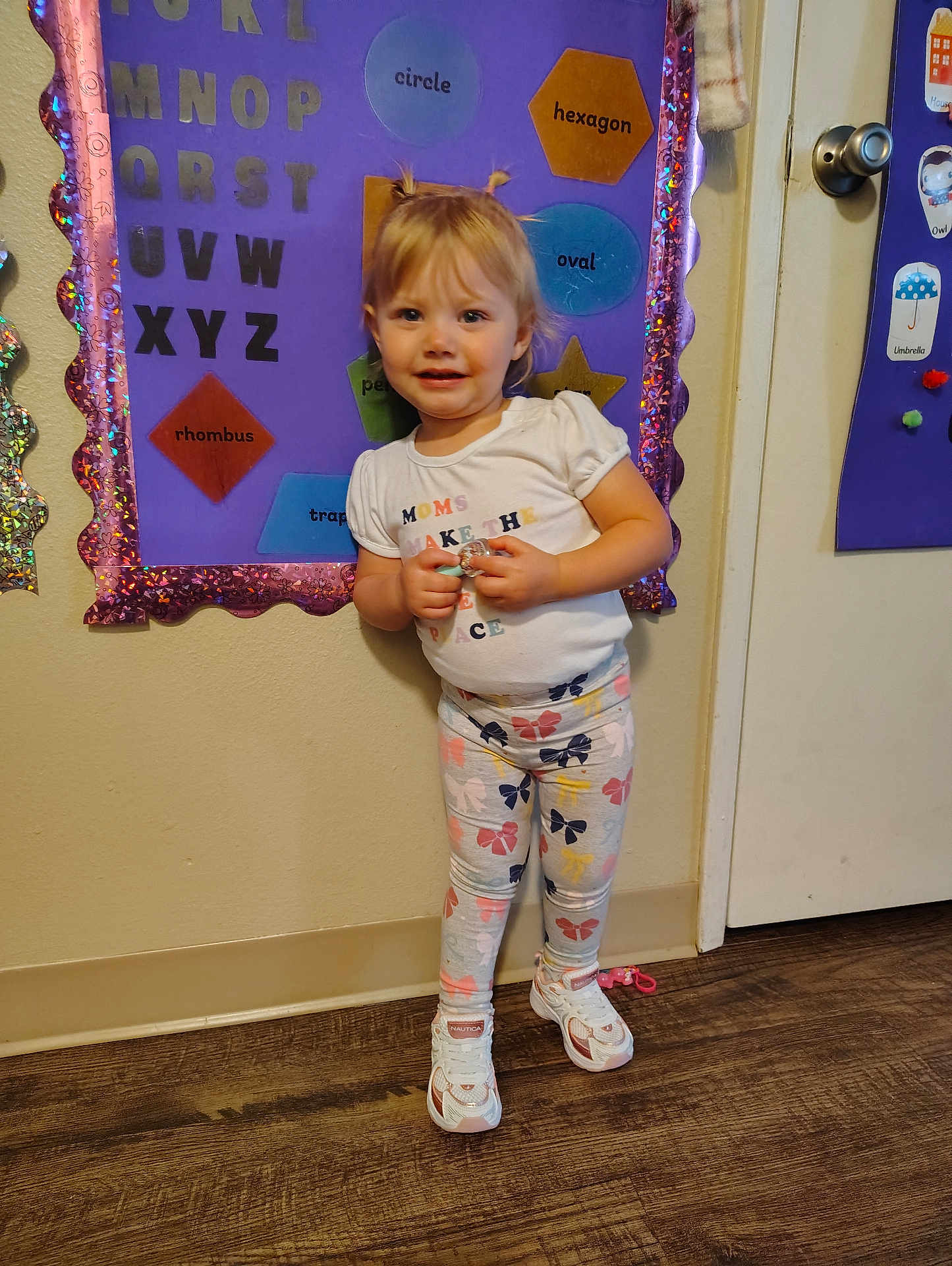 Charley is registered to the contest to win money with this photo: toddler, child, indoor, standing, smiling, leggings, sneakers, hair, pigtails, educational, bulletin_board, shapes, letters, floor, door, wall, clothing, person, cute, playful