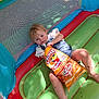 Charley is registered to the contest to win money with this photo: toddler, child, inflatable, playground, snack, puffcorn, cheese, food, bag, casual, relaxed, colorful, blue, red, green, yellow, smudges, face, legs, summer