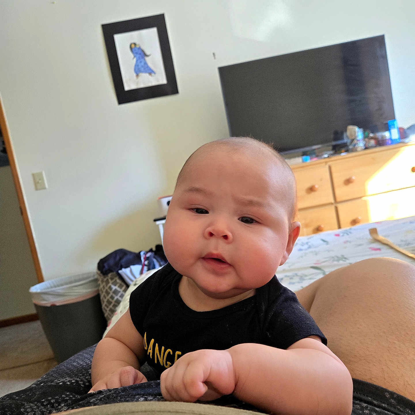 Josue is registered to the contest to win money with this photo: baby, infant, child, face, person, black_shirt, indoor, furniture, dresser, television, picture_frame, wall, bed, blanket, hand, skin, curious, seated, living_room, home