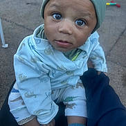 Amir is registered to the contest to win money with this photo: toddler, child, baby, face, eyes, beanie, hat, pajamas, dinosaur_pattern, socks, outdoor, person, hand, clothing, expression, curious, sitting, lap, ground, casual