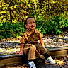 child, smiling, railroad_track, autumn_leaves, forest, outdoor, greenery, sunlight, casual_clothing, white_sneakers, nature, daylight, happy, person, seasonal, trees, wooden_planks, fall, young_child, sitting