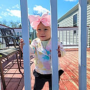 Catalina is registered to the contest to win money with this photo: toddler, child, pink_bow, colorful_jacket, black_pants, porch, railing, outdoor, sunny, blue_sky, wooden_deck, smile, person, daylight, playful, hand, face, clothing, baby, young_child