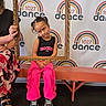 adult_hand, background, bench, black_top, casual_clothing, child, dance, event, expression, frame, girl, hair_tied, indoor, logo, pink_pants, playful, portrait, sign, sneakers, tongue_out