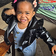 Aëla a rejoint le concours — aidez-le/la à gagner de superbes lots ! toddler, child, smiling, leather_jacket, denim_dress, hair_clips, indoor, carpet, table, cushion, floor, playful, person, cute, happy, portrait, clothing, footwear, furniture, home