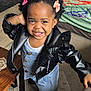 toddler, child, smiling, leather_jacket, denim_dress, hair_clips, indoor, carpet, table, cushion, floor, playful, person, cute, happy, portrait, clothing, footwear, furniture, home