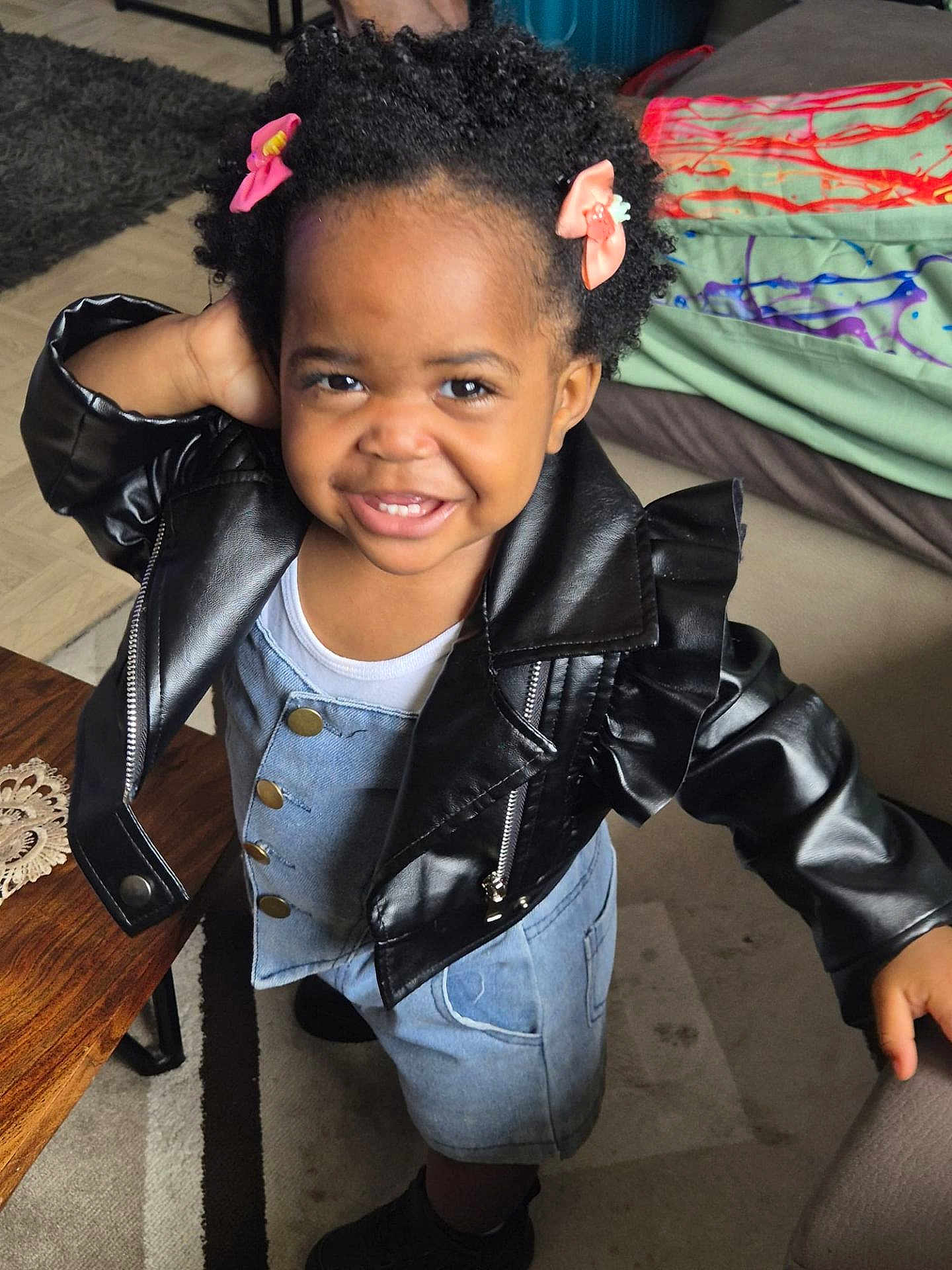 Aëla a rejoint le concours — aidez-le/la à gagner de superbes lots ! toddler, child, smiling, leather_jacket, denim_dress, hair_clips, indoor, carpet, table, cushion, floor, playful, person, cute, happy, portrait, clothing, footwear, furniture, home
