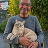 animal, casual_clothing, cat, chair, cream_colored_cat, daylight, glasses, greenery, grey_sweater, hands, happy, man, nature, older_adult, outdoor, pet, portrait, relaxed, sitting, smiling