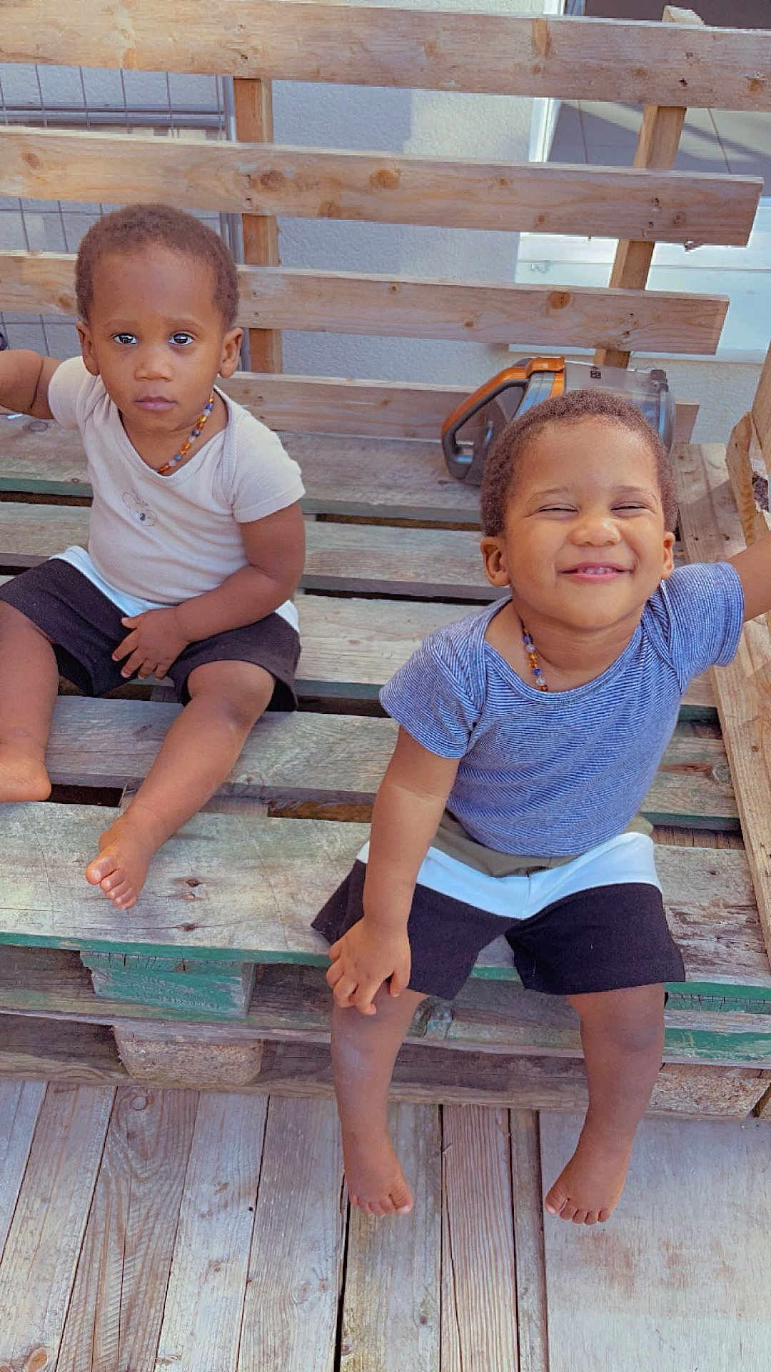 Ousmane participe au concours pour gagner de l'argent avec cette photo : child, toddler, smiling, serious, barefoot, wooden_bench, outdoor, casual_clothing, necklace, shorts, short_sleeve_shirt, wooden_floor, pallet, two_children, happy, cute, playful, sitting, friends, daylight