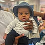 Dakota is registered to the contest to win money with this photo: baby, infant, child, hat, bucket_hat, eyes, face, hands, adult_hand, holding, white_shirt, black_pants, socks, indoor, store, blurred_background, portrait, stroller, mobile_screenshot, social_media_ui