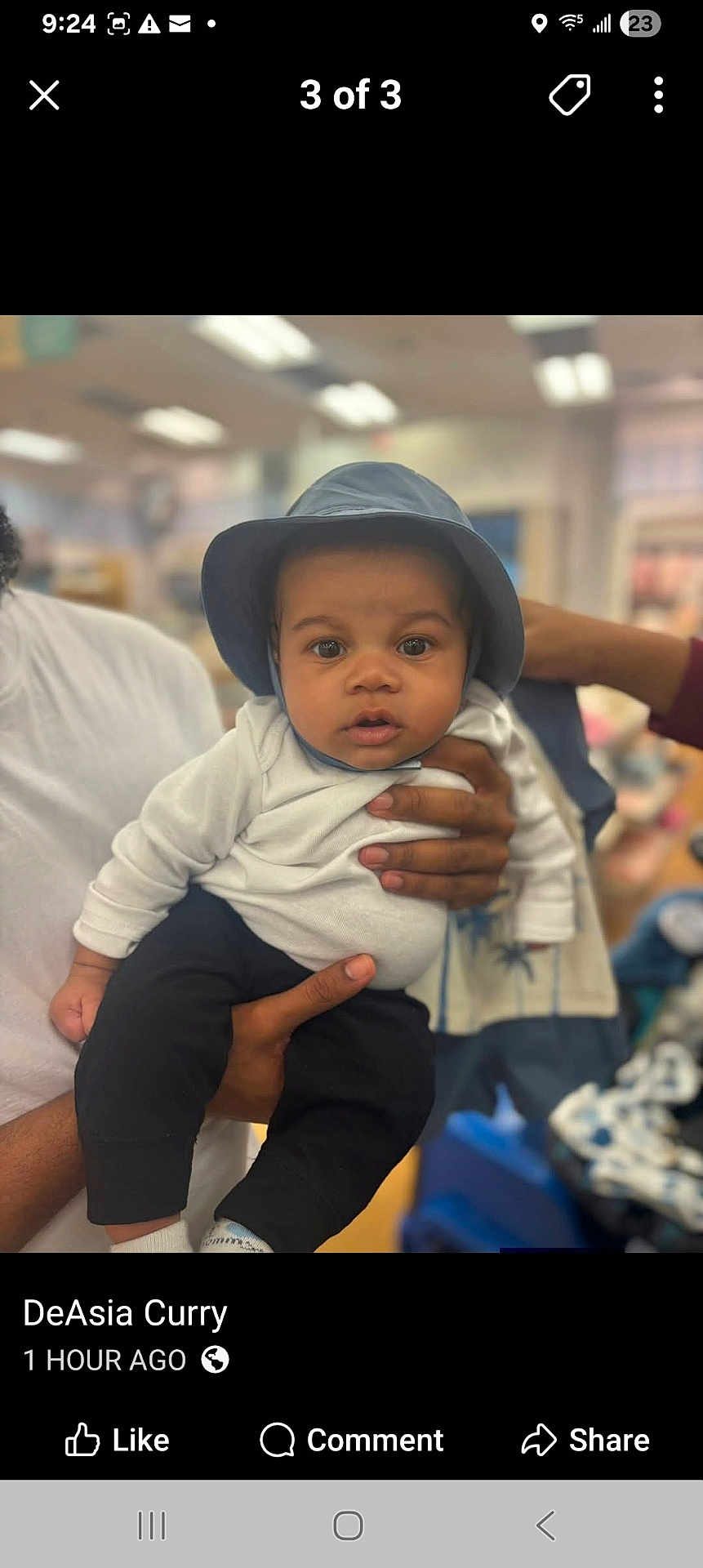 Dakota is registered to the contest to win money with this photo: baby, infant, child, hat, bucket_hat, eyes, face, hands, adult_hand, holding, white_shirt, black_pants, socks, indoor, store, blurred_background, portrait, stroller, mobile_screenshot, social_media_ui