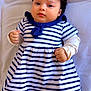baby, infant, child, curly_hair, striped_dress, navy_blue, white_clothing, lying_down, bed, soft_fabric, cute, small_fists, portrait, indoors, calm_expression, young_child, closeup, person, sleepwear, adorable