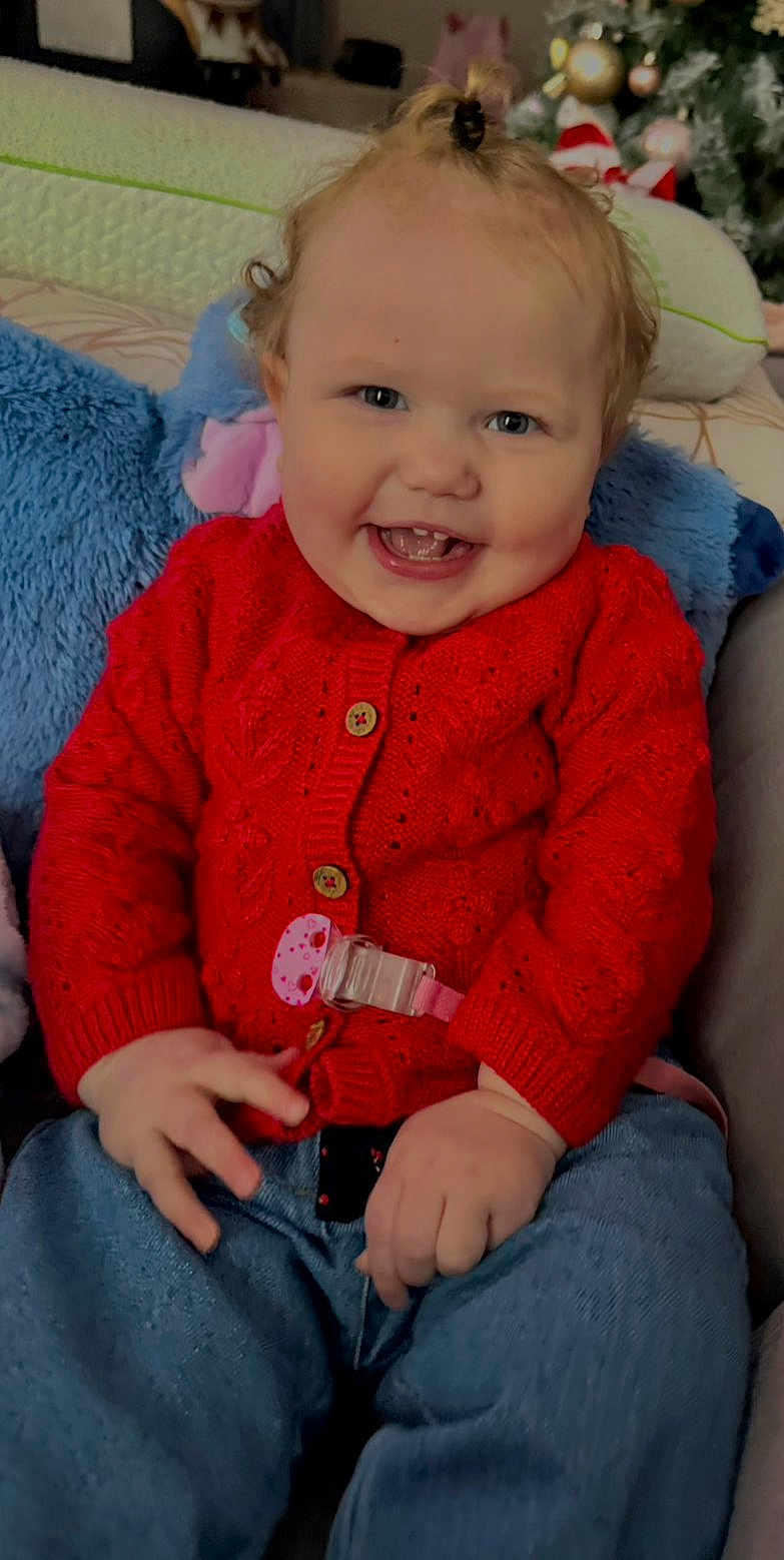 Ambre a rejoint le concours — aidez-le/la à gagner de superbes lots ! baby, child, smiling, red_sweater, blue_jeans, indoor, face, happy, cute, plush_toys, couch, portrait, sitting, human, clothing, hand, hair, holiday_decor, tooth, pacifier_clip