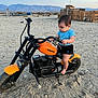 Michelle is registered to the contest to win money with this photo: toddler, child, motorcycle, toy, orange, black, sand, desert, mountains, fence, blue_shirt, shorts, outdoor, vehicle, baby, curious, play, handlebars, sunset, foot