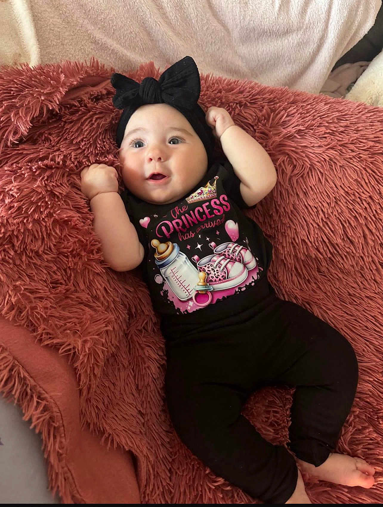 Lina participe au concours pour gagner de l'argent avec cette photo : baby, infant, black_clothing, headband, bow, blanket, pacifier_graphic, baby_bottle_graphic, pink_text, cute, smiling, lying_down, cozy, soft_texture, child, young, happy, indoors, person, small_feet