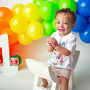 Zaylin is registered to the contest to win money with this photo: toddler, child, birthday, first_birthday, balloons, rainbow, toy, white_chair, smiling, curly_hair, clapping_hands, celebration, colorful, happy, indoor, number_one, shorts, tshirt, studio, portrait