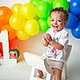 toddler, child, birthday, first_birthday, balloons, rainbow, toy, white_chair, smiling, curly_hair, clapping_hands, celebration, colorful, happy, indoor, number_one, shorts, tshirt, studio, portrait